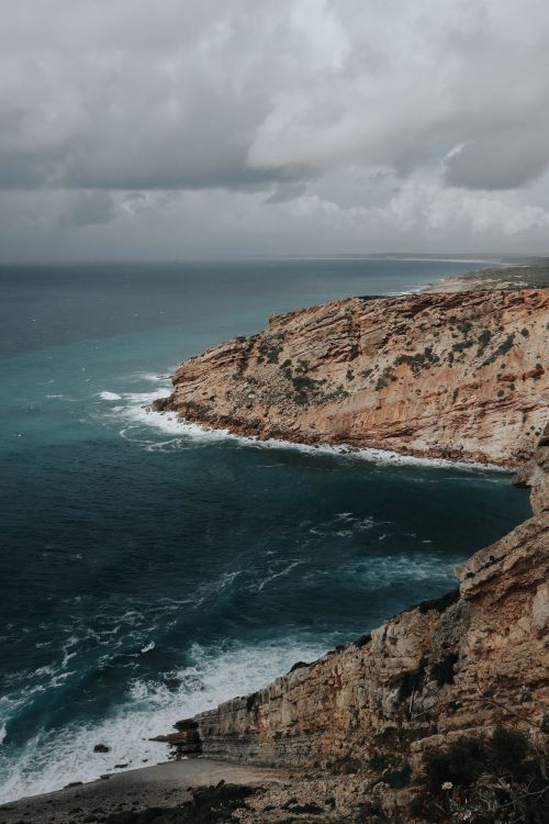Free Stunning view of rugged cliffs meeting the ocean under a dramatic cloudy sky. Stock Photo