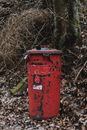 Red shabby trash can placed in autumn park with yellow fallen leaves and dry trees