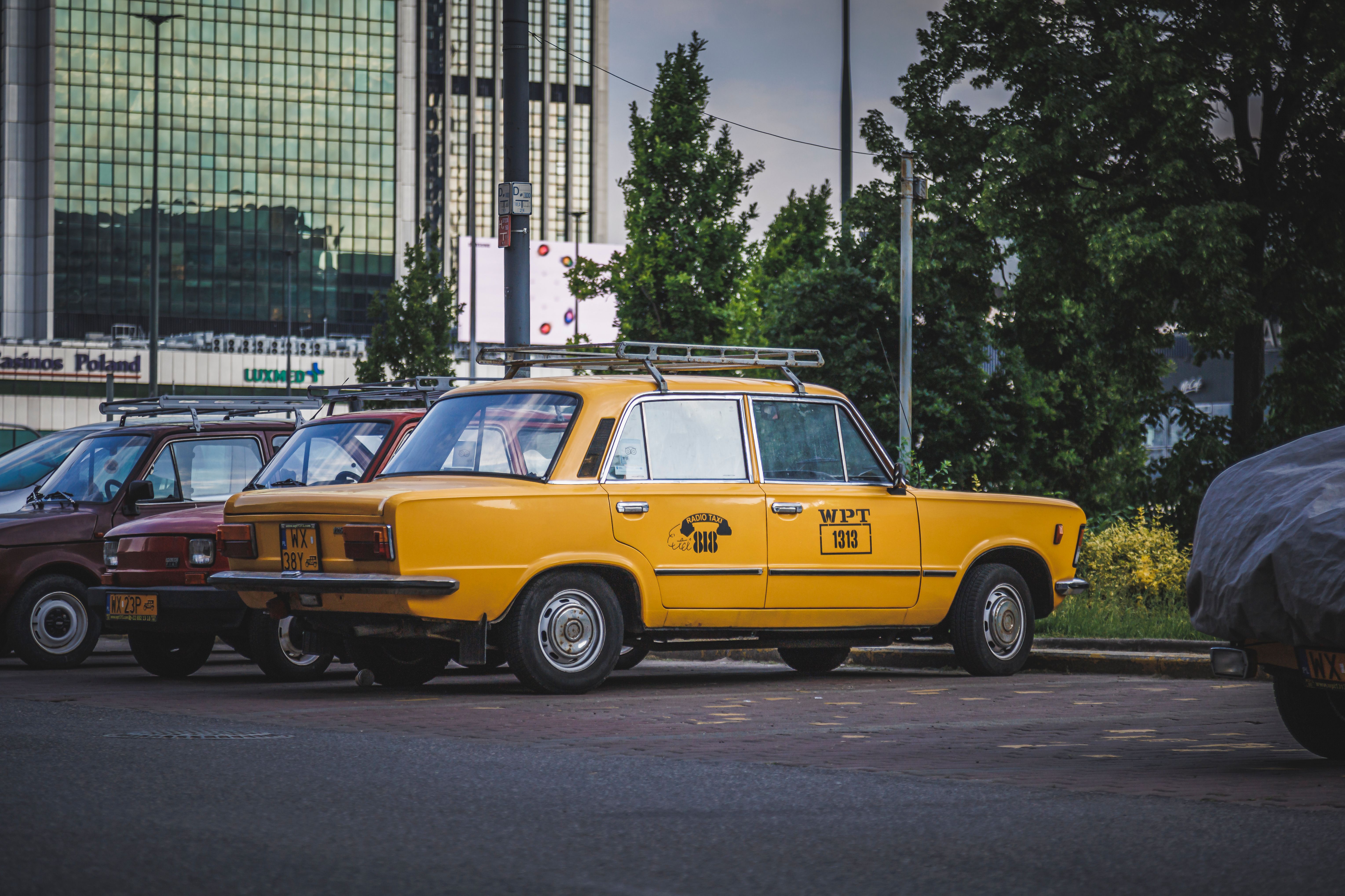 Classic yellow car parked in a city lot with a backdrop of buildings and greenery.