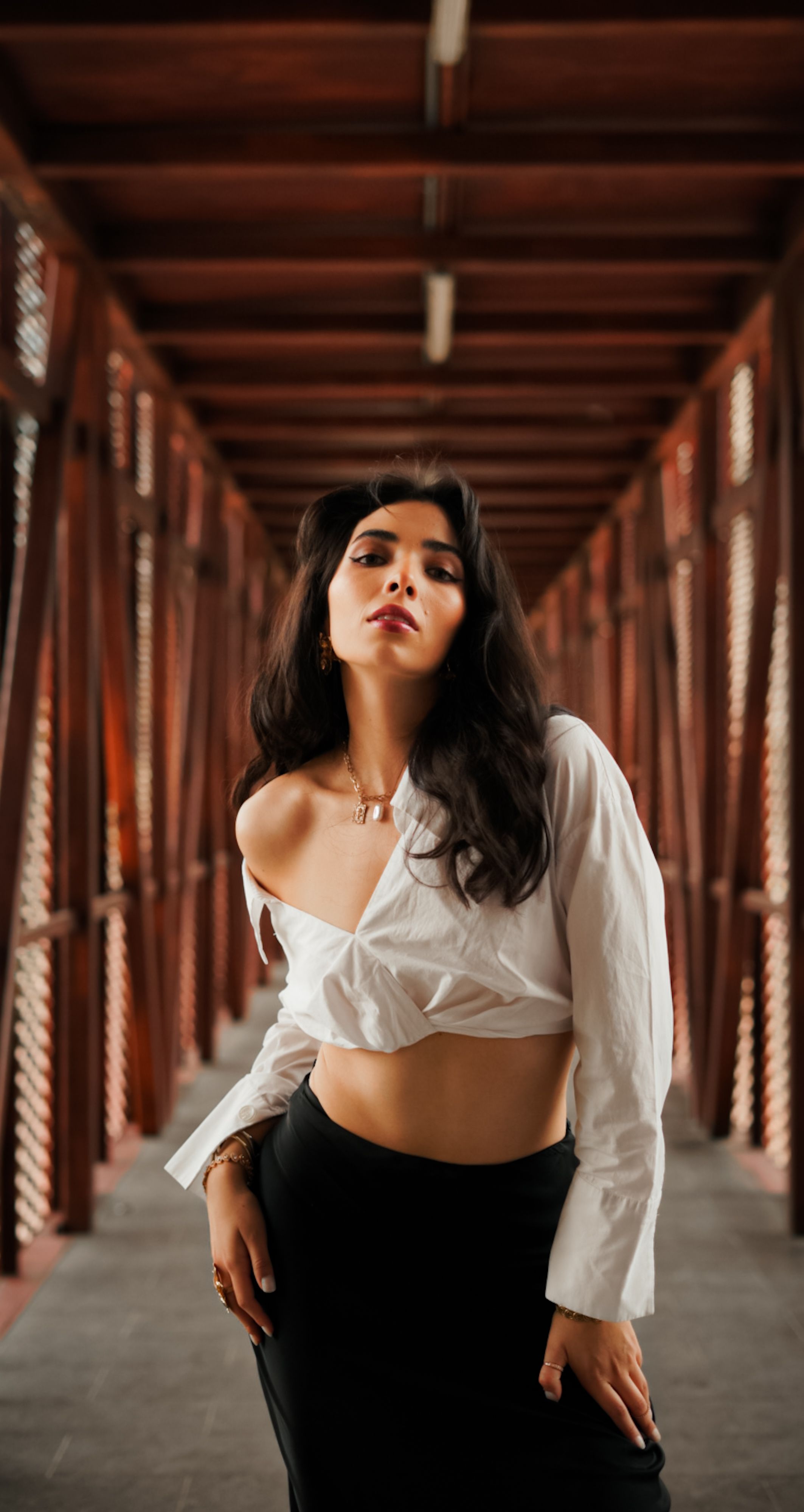 A fashionable woman poses confidently in a modern wooden bridge, wearing a white blouse and black skirt.
