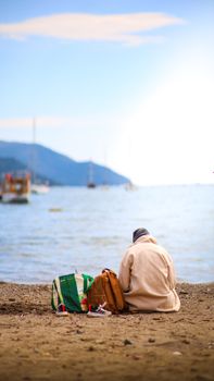 Person sitting alone on a sandy beach with calm sea and distant boats.