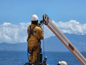 A dock worker in yellow uniform operates machinery against a stunning sea and mountain backdrop.