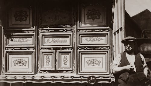 Grayscale Photo of Man Standing Beside A Wooden Wall With Carvings