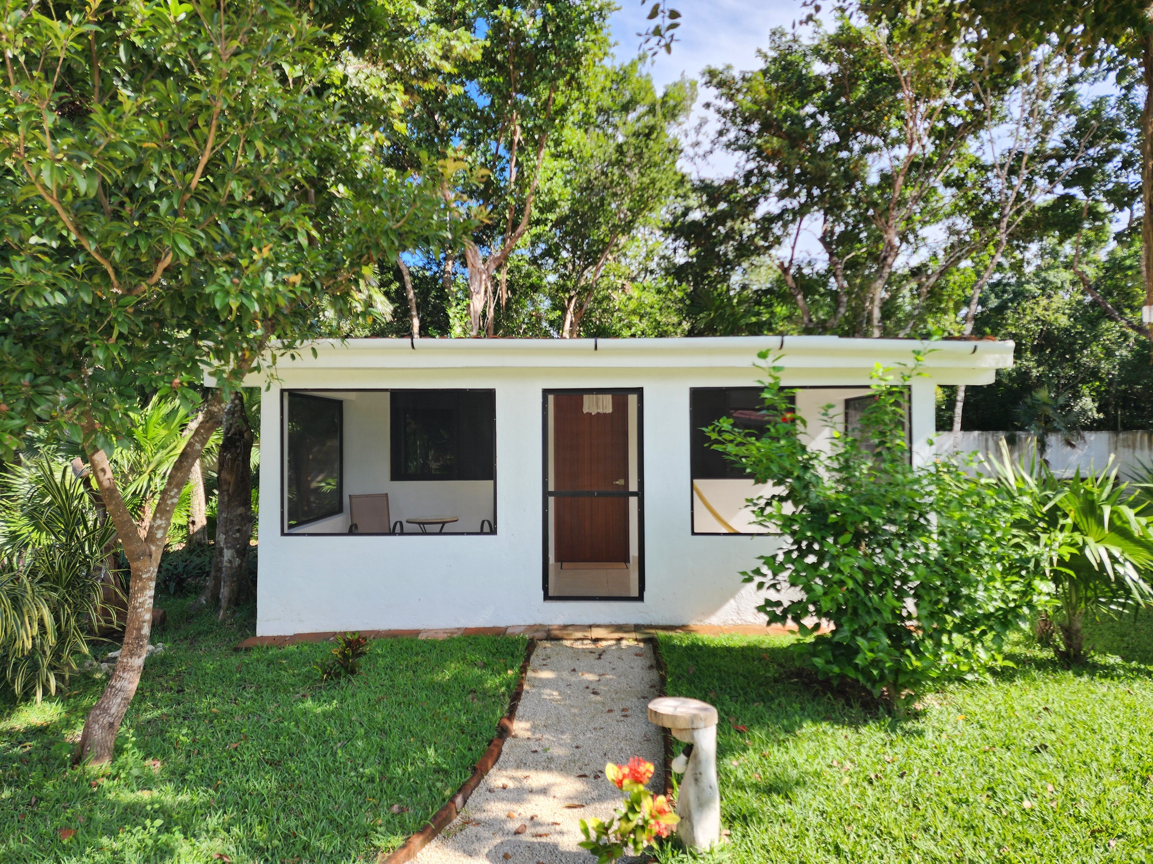 Small bungalow in a lush garden setting with green grass and trees under clear blue sky.