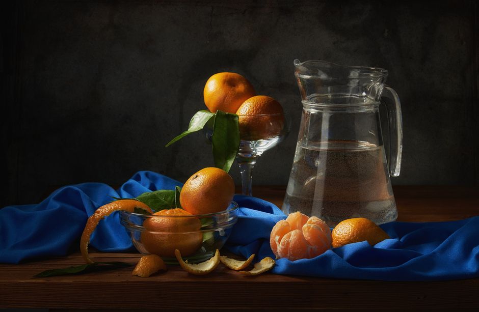 A still life composition featuring tangerines on a blue cloth beside a glass jug of water.