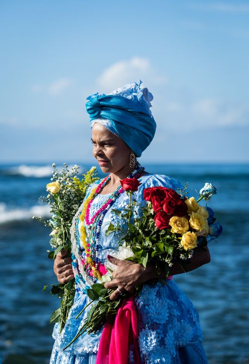 Free Vibrant portrait of an African woman in traditional attire holding flowers by the sea in Salvador, Brazil. Stock Photo