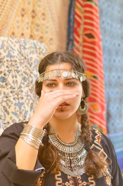 Free A woman adorned with ethnic jewelry and headpiece, posing indoors by colorful rugs. Stock Photo