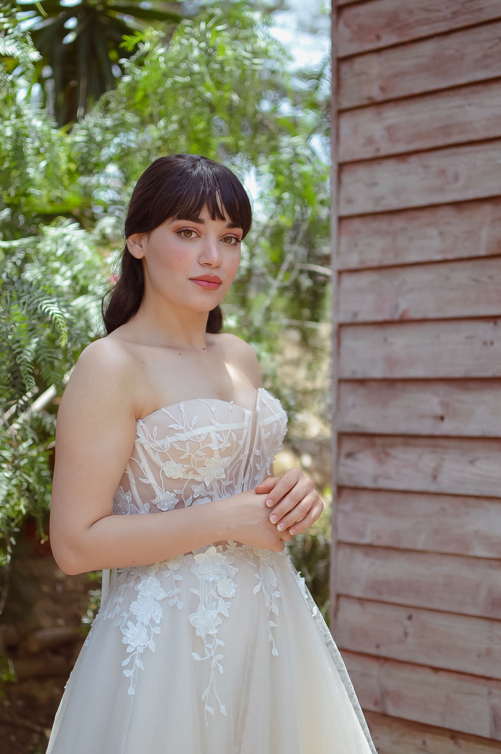 A beautiful bride in a lace wedding dress poses by a wooden structure surrounded by greenery.