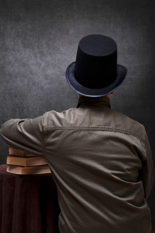 Free A man wearing a top hat sits by a stack of books, facing away against a dark backdrop. Stock Photo