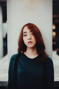 一位红发、表情严肃、身着休闲服的年轻亚洲女性站在白柱附近看着镜头