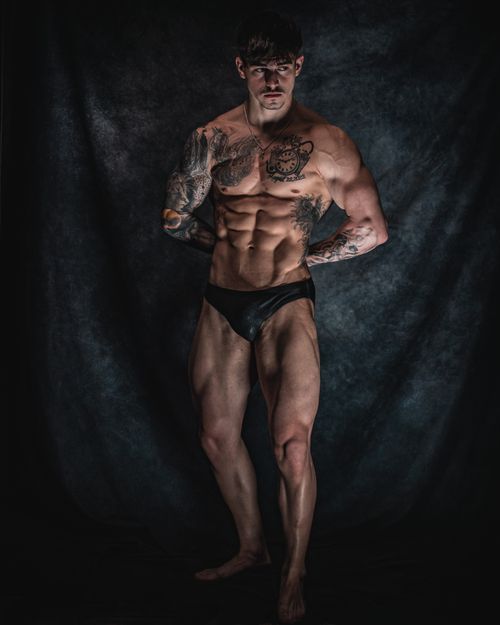 Free A muscular male model poses confidently in dark studio lighting, showcasing tattoos and physique. Stock Photo