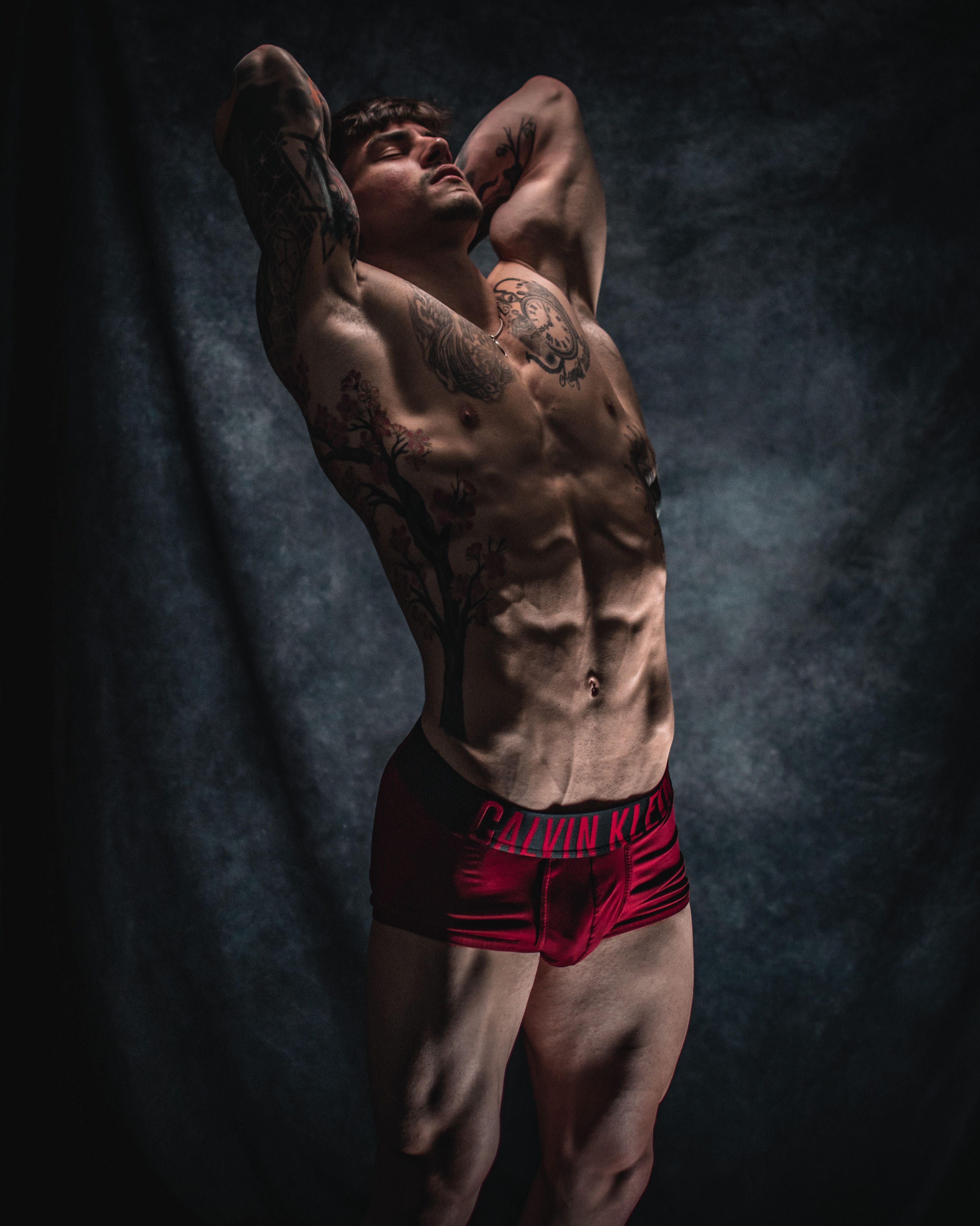 Dramatic studio shot of a tattooed male model posing in red underwear under moody lighting.