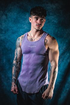 Strong, tattooed man in a purple tank top against a moody blue background.