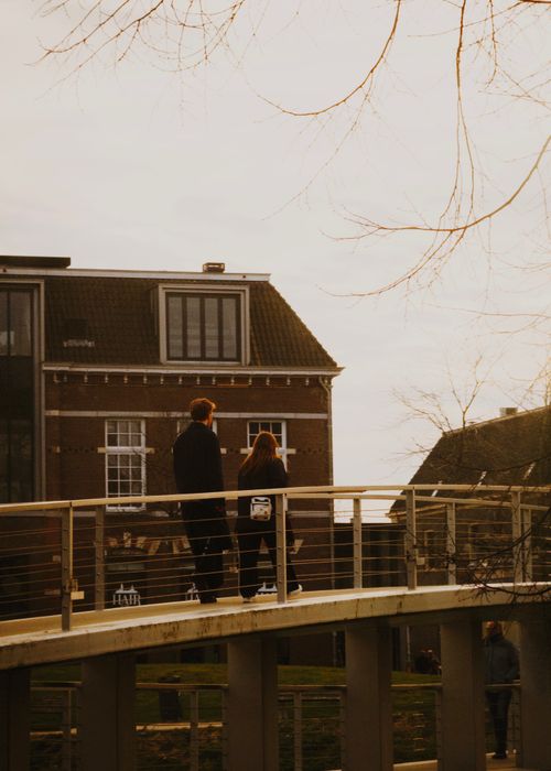 Free A couple walking on a modern bridge with traditional Dutch buildings in the background at sunset. Stock Photo