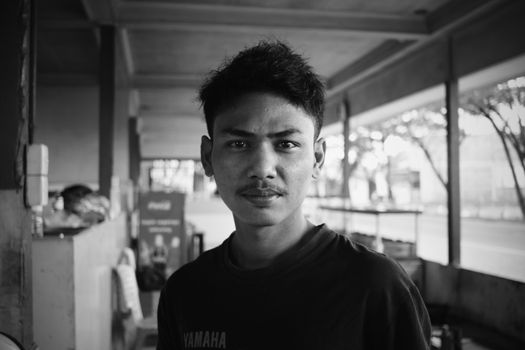 Black and white of young unshaven Asian guy standing in cafe and looking at camera