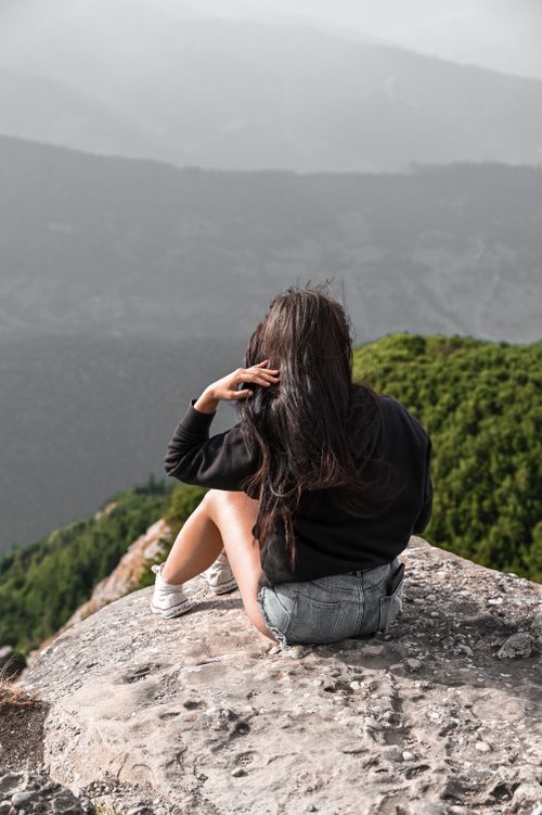 Free Woman sitting on rocky cliff edge with scenic mountain view, embodying peace and nature connection. Stock Photo