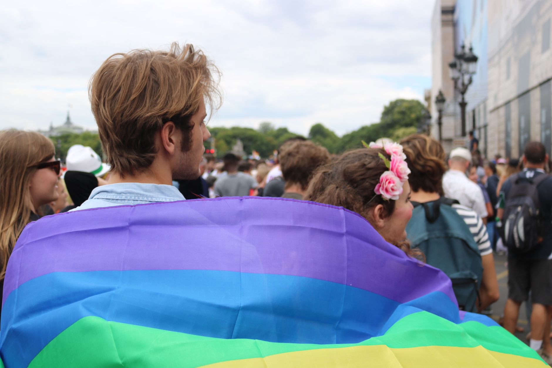 Młodzi ludzie widziani od tyłu, owinięci tęczową flagą podczas marszu równości w mieście
