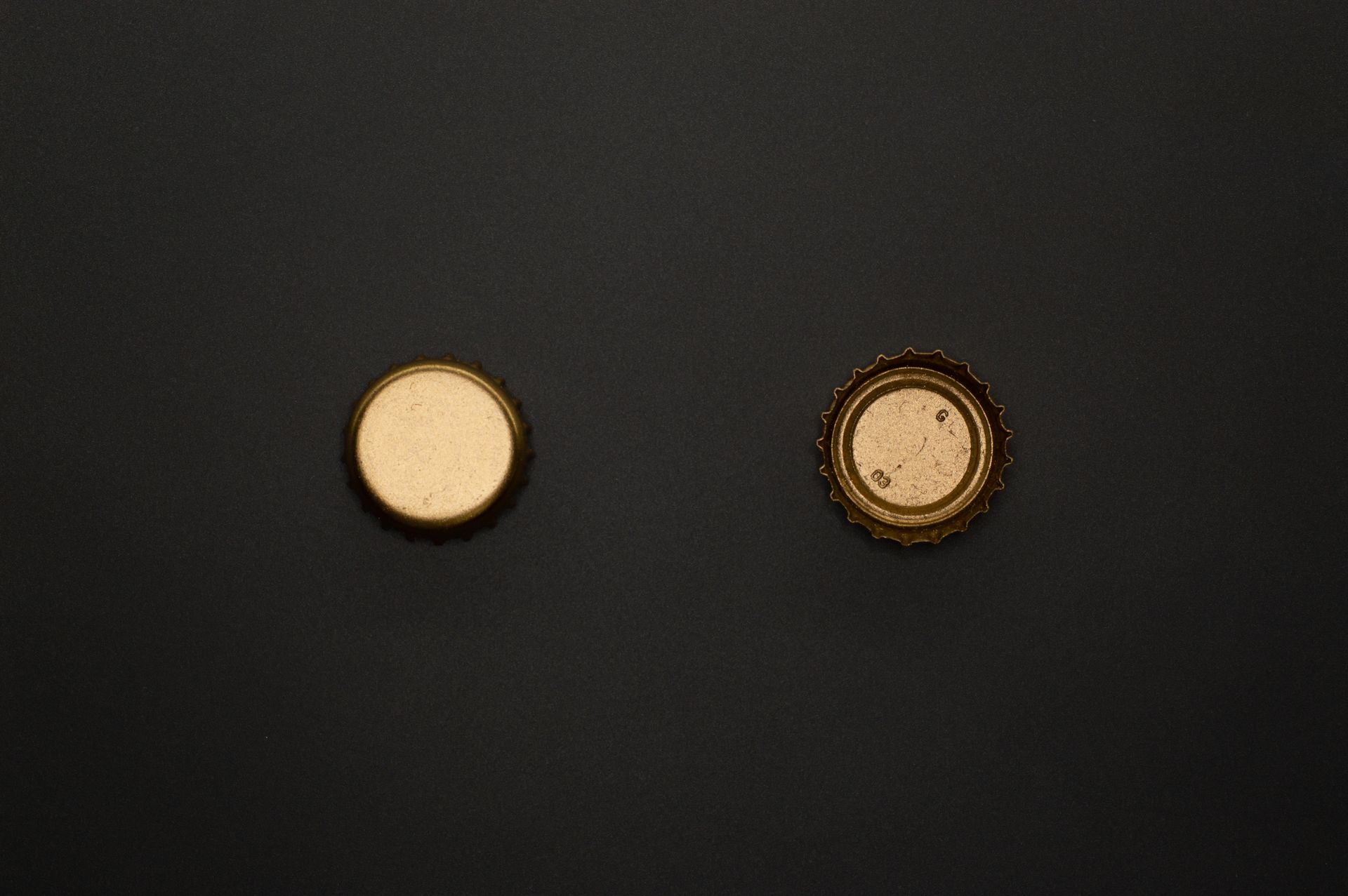 Minimalistic photo of two golden beer caps against a dark background, highlighting texture and color contrast.