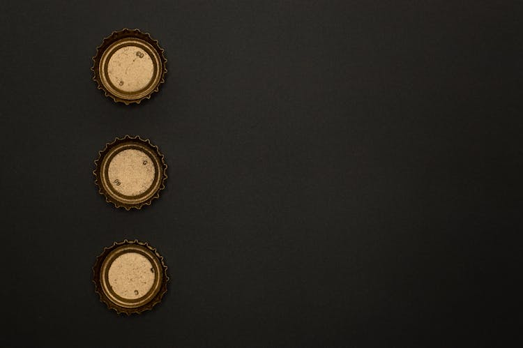 Three Gold Round Bottle Tops On Black Surface