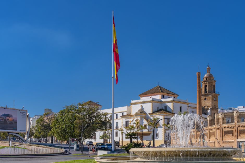 Vista escénica de una plaza en Cádiz con una destacada bandera española, una fuente y arquitectura histórica.