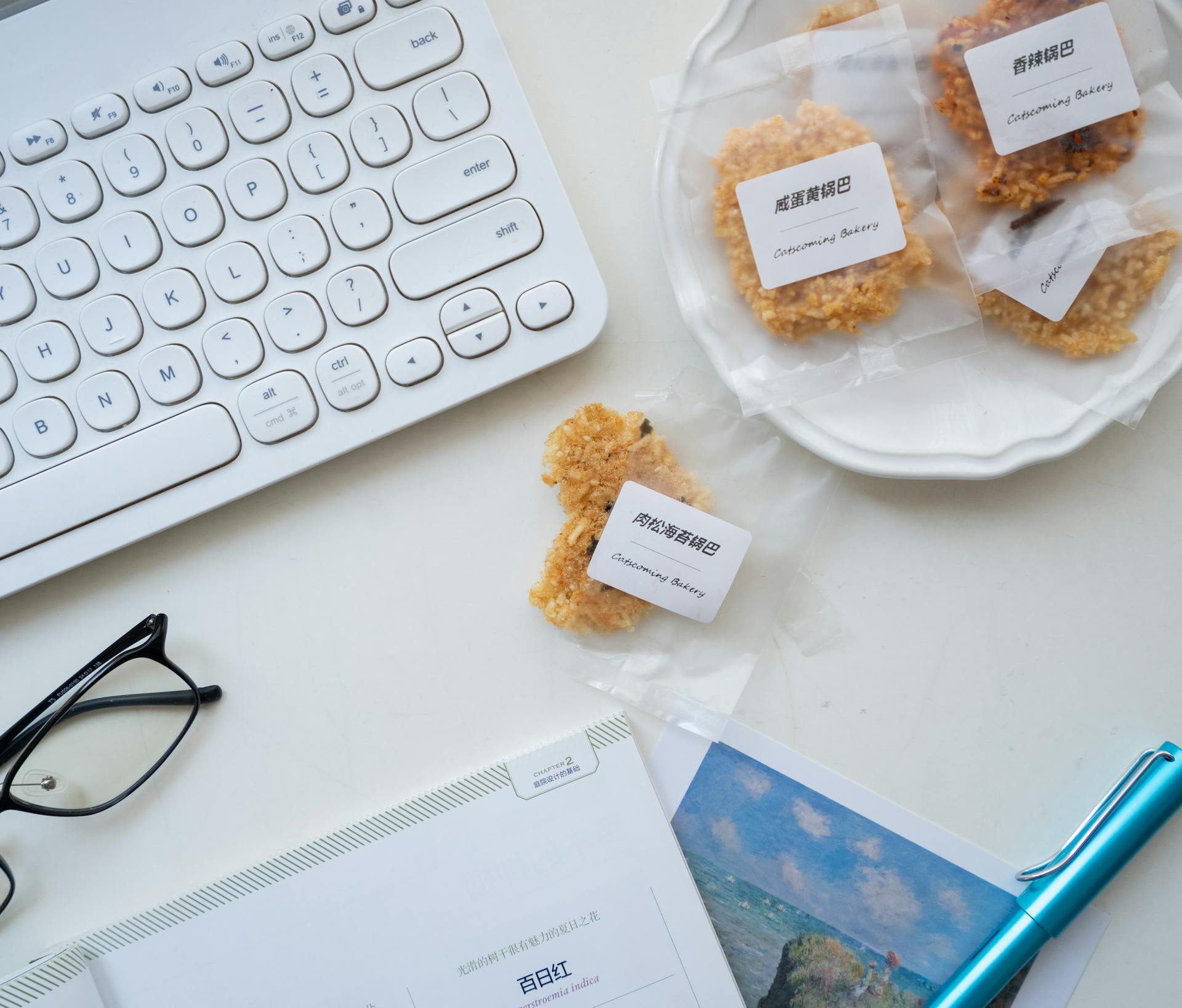 Top view of a workspace with a keyboard, wrapped snacks, glasses, and a notebook.