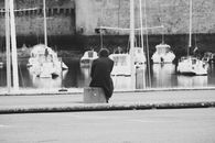 A lone person sits contemplatively by a harbor, watching the boats in a calm atmosphere.