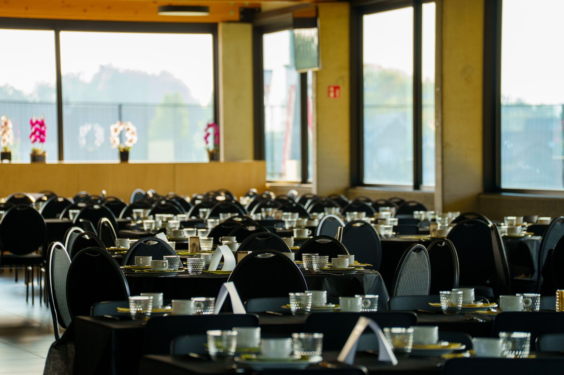 Spacious dining hall set for an event with black table settings and large windows.