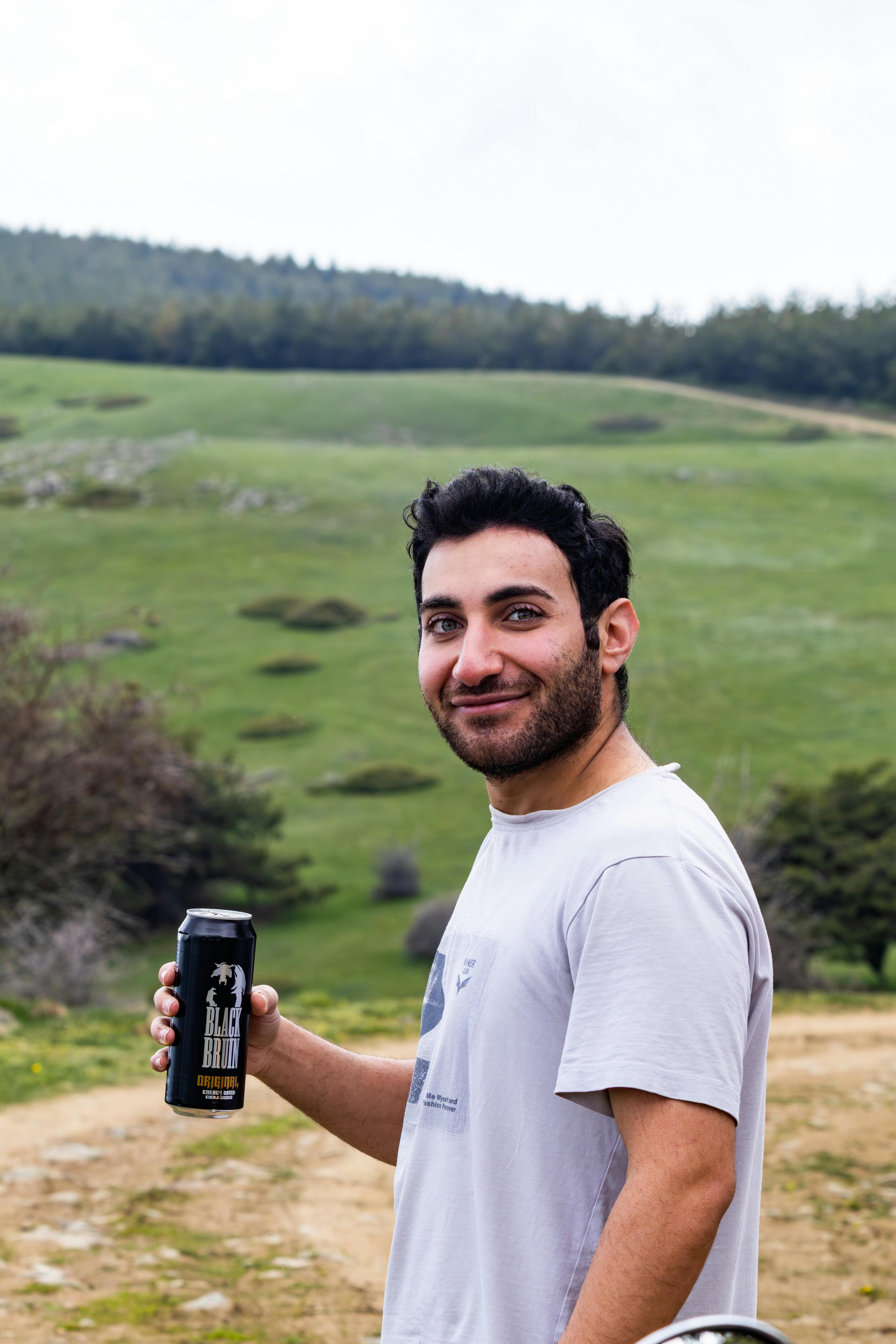 A smiling man holding a drink can in a lush, green outdoor setting.