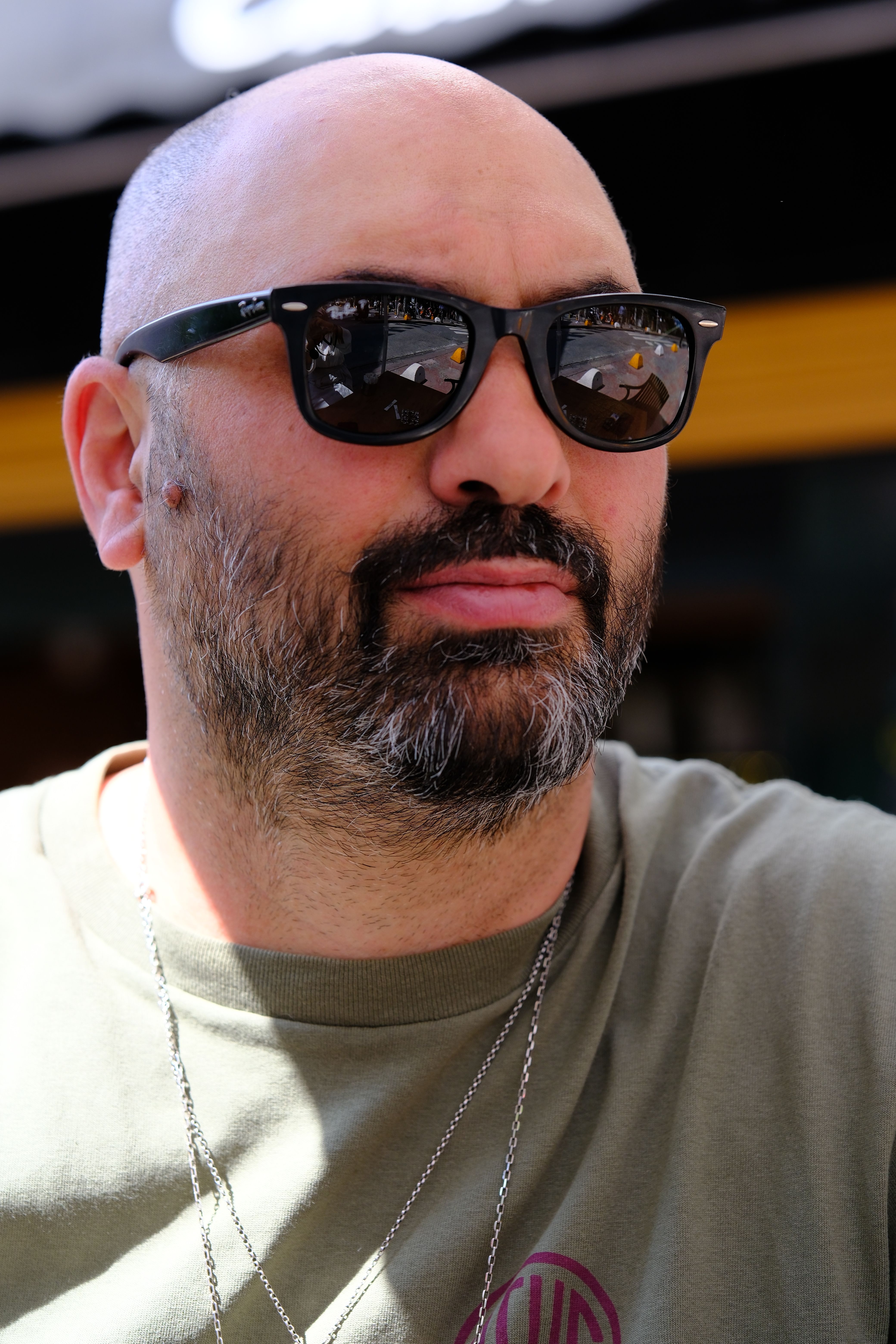 A bald man with sunglasses sitting indoors, reflecting a casual vibe.