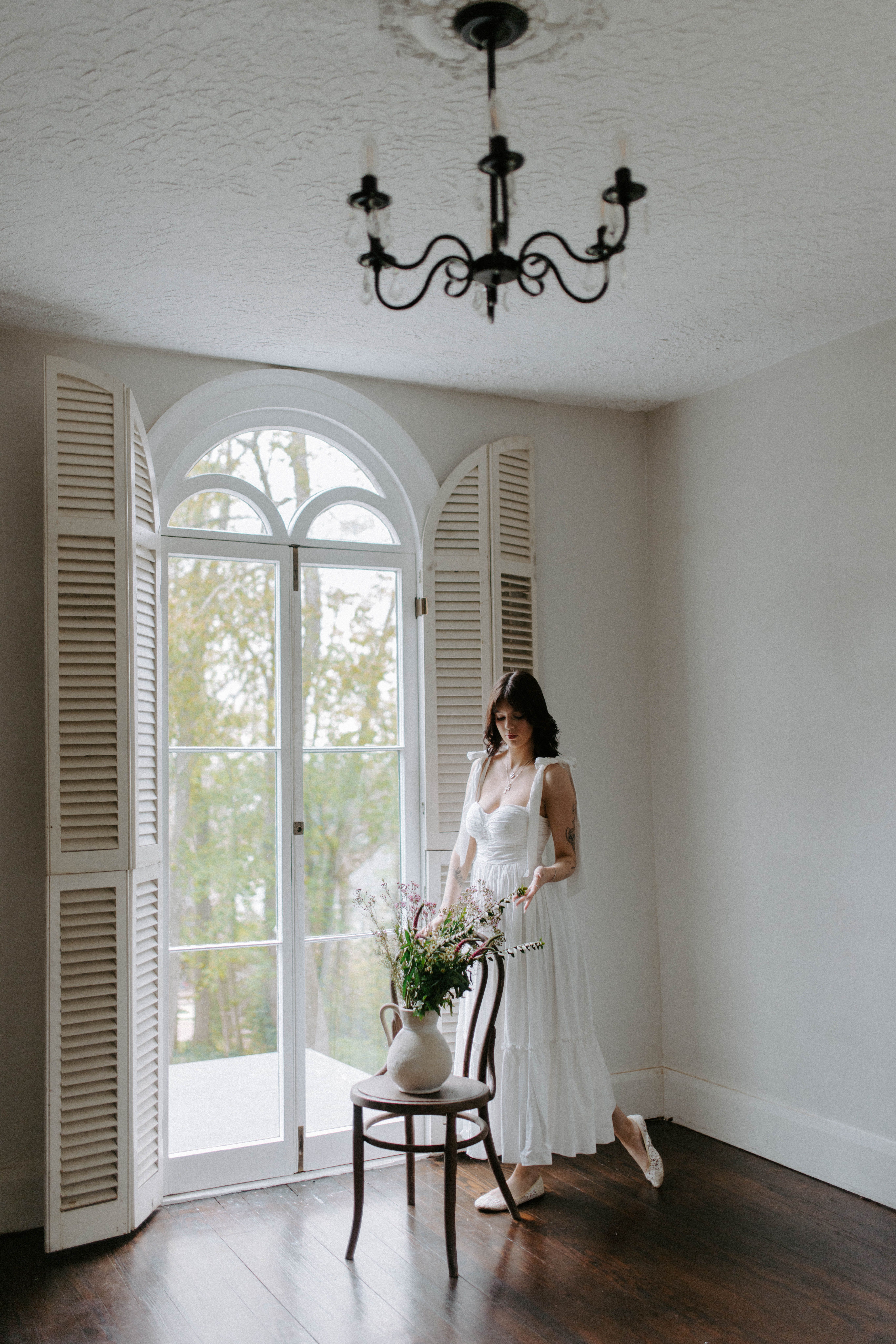 Woman in flowing dress by arched window in minimalist room with floral arrangement.