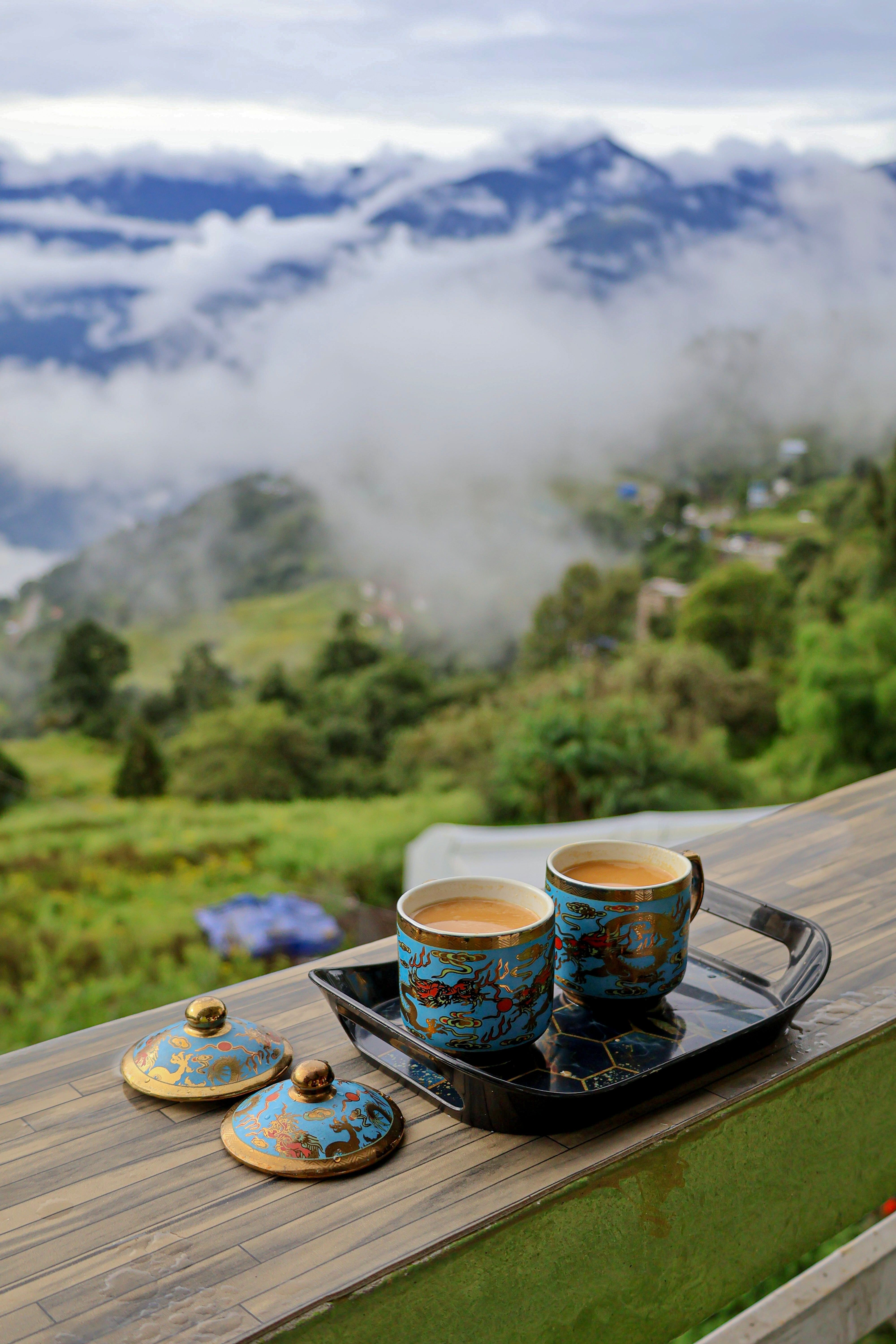 Dos tazas de té con tapa, azules y doradas, en una bandeja sobre la barandilla de una veranda de madera, con el valle lleno de nubes