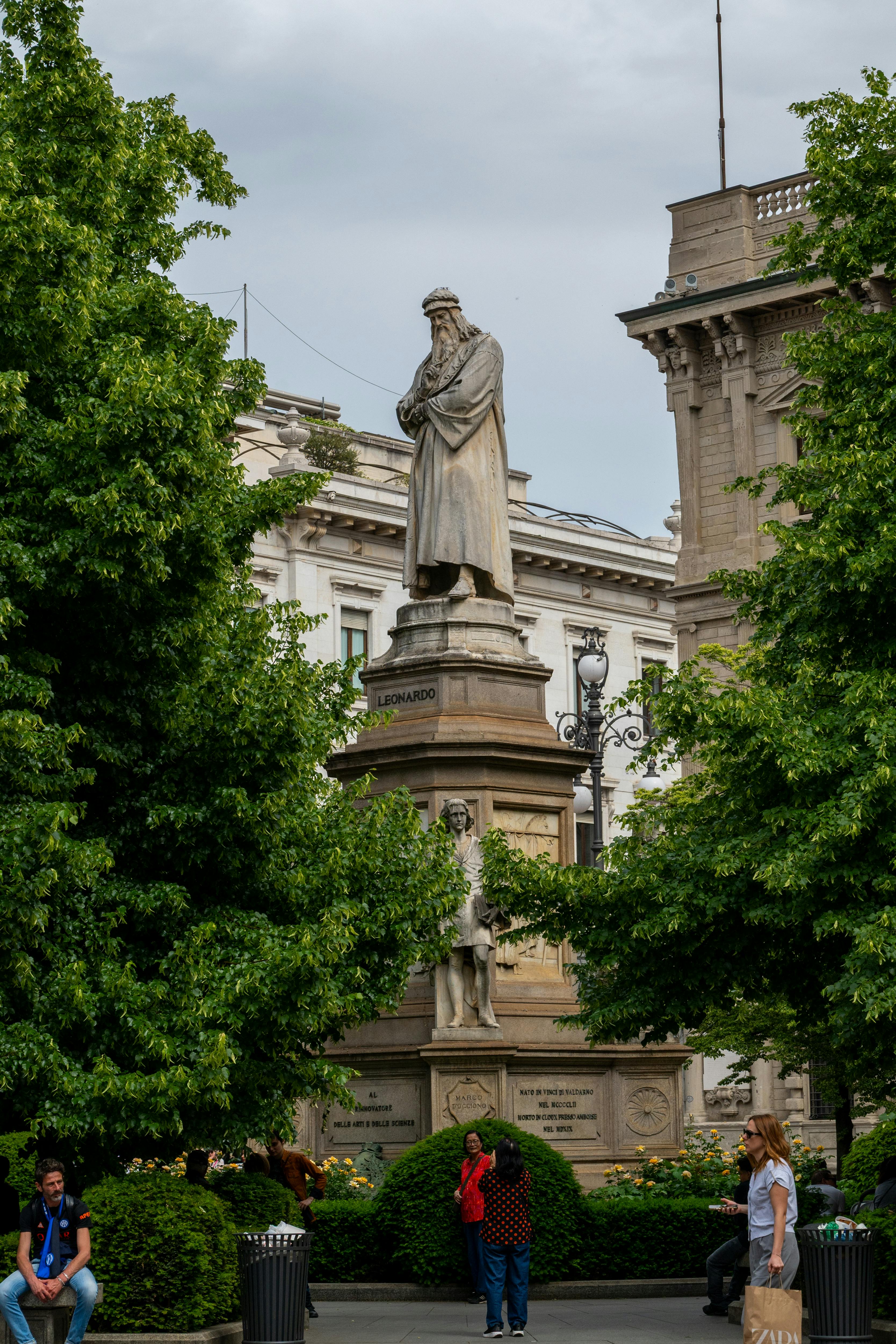 Statue of Leonardo da Vinci located in Milan surrounded by greenery and people.