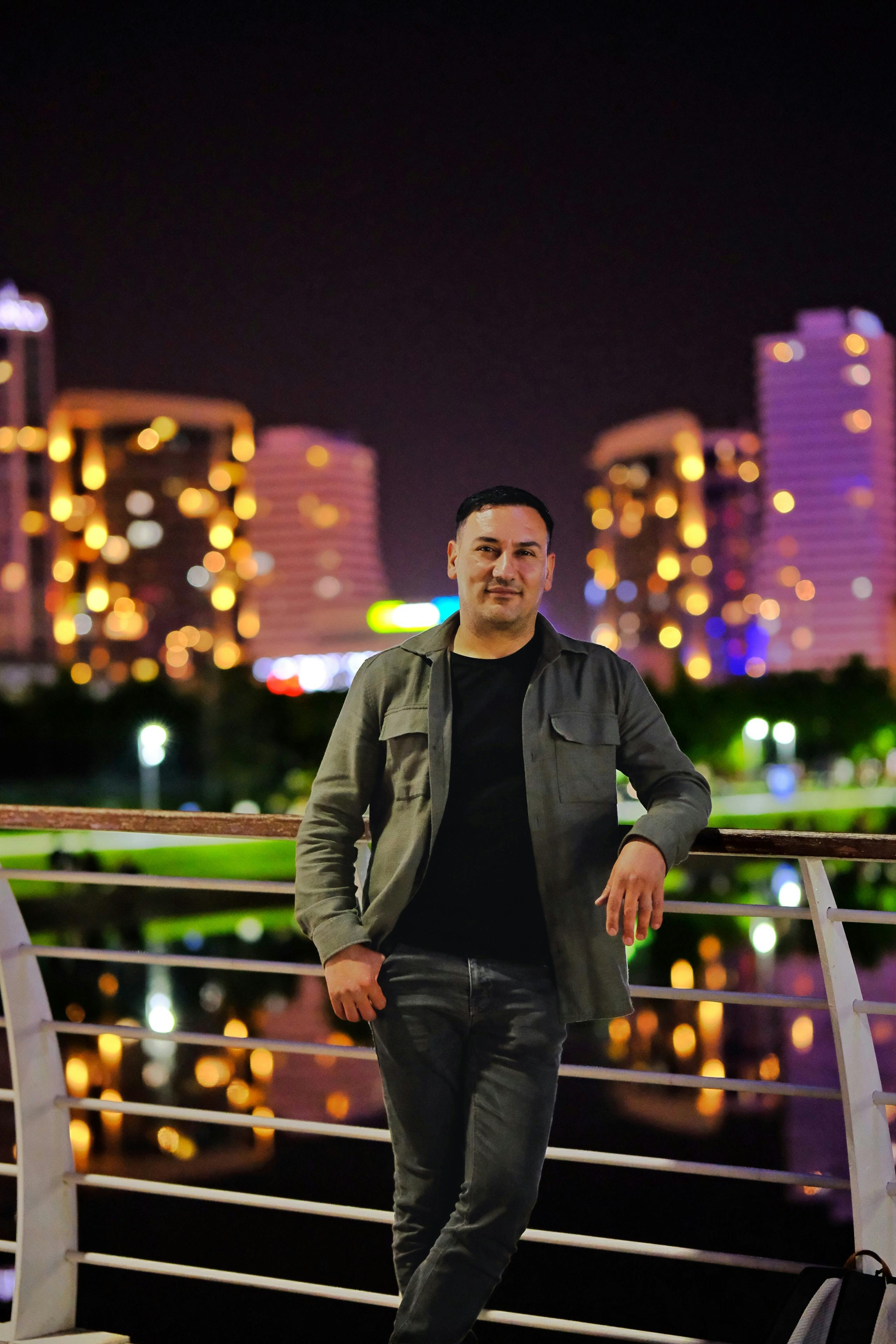 Nighttime portrait of a man on a bridge with city skyline lights in the background.