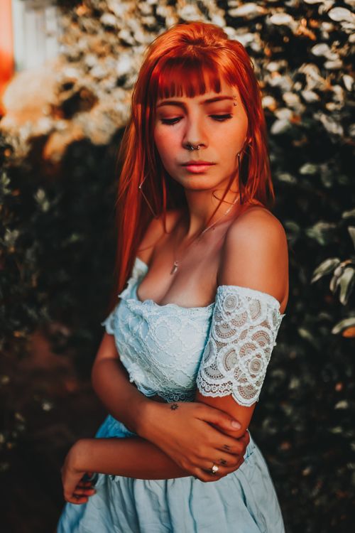 Free Young lady with long ginger hair in white stylish dress with bare shoulders standing near green bushes and looking down while resting in garden on summer sunny day Stock Photo