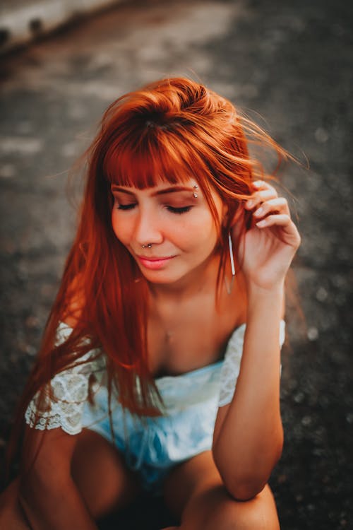 Free From above of slim young sensual female with closed eyes and eyebrow piercing in blouse and earring squatting while touching red hair outdoors Stock Photo