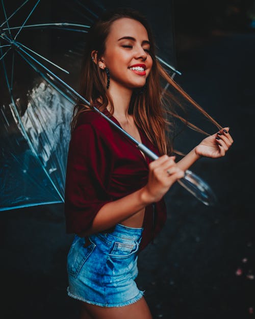 Side view of young fit positive female with closed eyes in denim shorts strolling with transparent umbrella while touching hair and strolling on black background