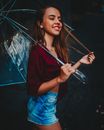 一位年轻健美的女性闭眼侧身，穿着牛仔短裤，撑着透明雨伞在黑色背景中漫步，同时轻抚头发