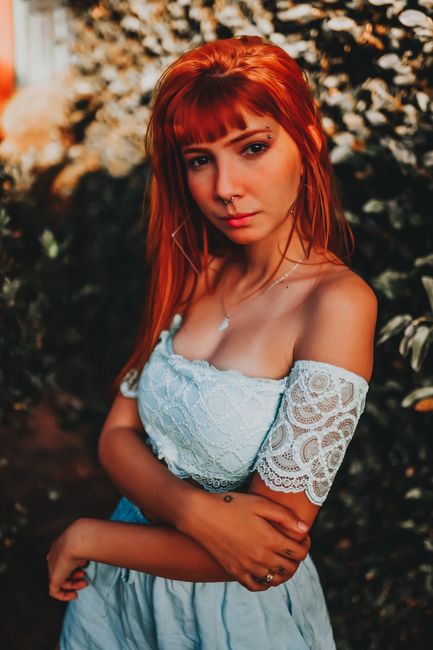 Thoughtful young slender female with red hair in elegant dress standing near tree with blossoming flowers and looking at camera in summer