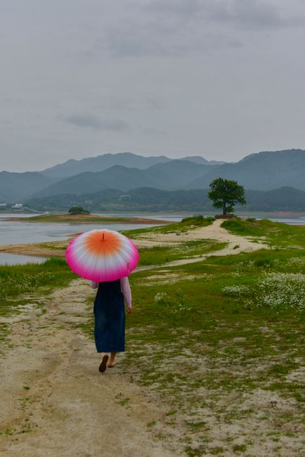 A woman with a colorful umbrella walks along a lakeside path surrounded by mountains and greenery.