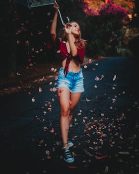 Woman posing outdoors with umbrella amidst falling leaves on a scenic road.