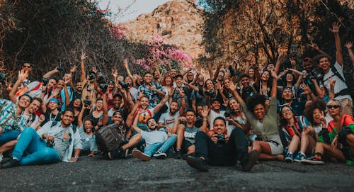 Free Group of multiracial happy people sitting with raised hands on asphalt road near mountains and trees in summer for portrait photo and looking at camera Stock Photo