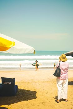 A sunny day at Bali beach with people surfing and relaxing under umbrellas.