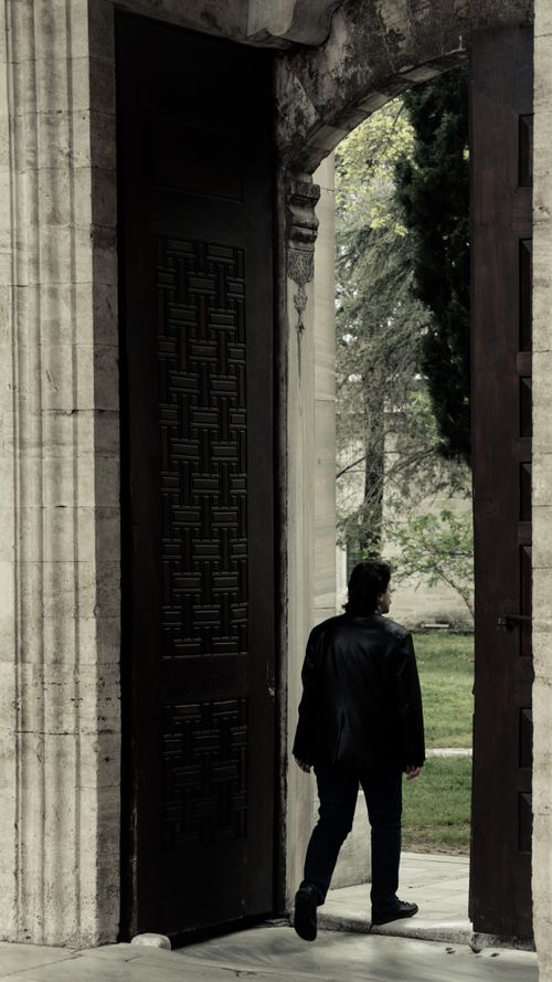 Free Dark-clothed man exits through a large historic stone archway into a lush green outdoor space. Stock Photo