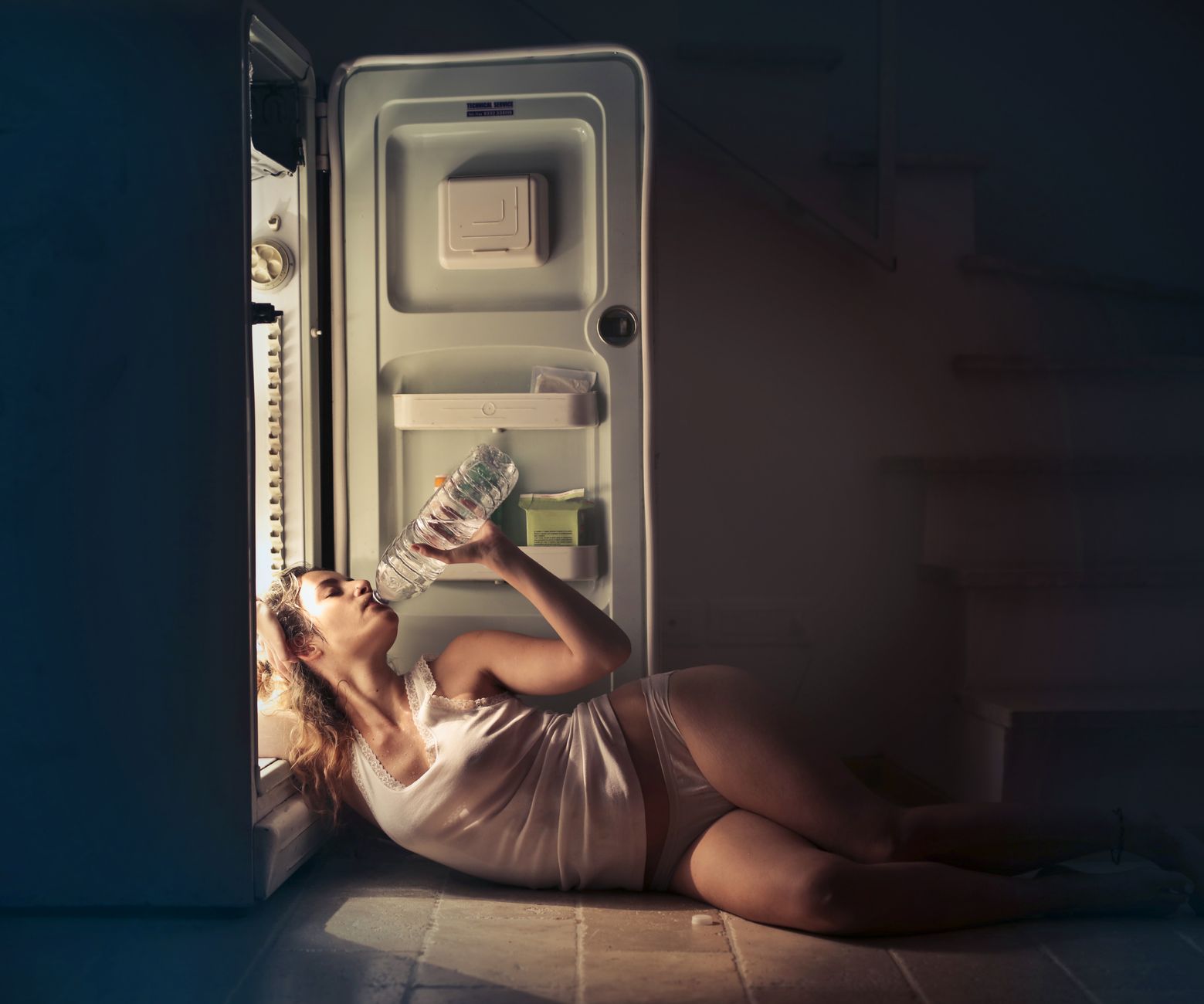 A woman in sleepwear drinking water beside an open fridge late at night, conveying thirst and refreshment.