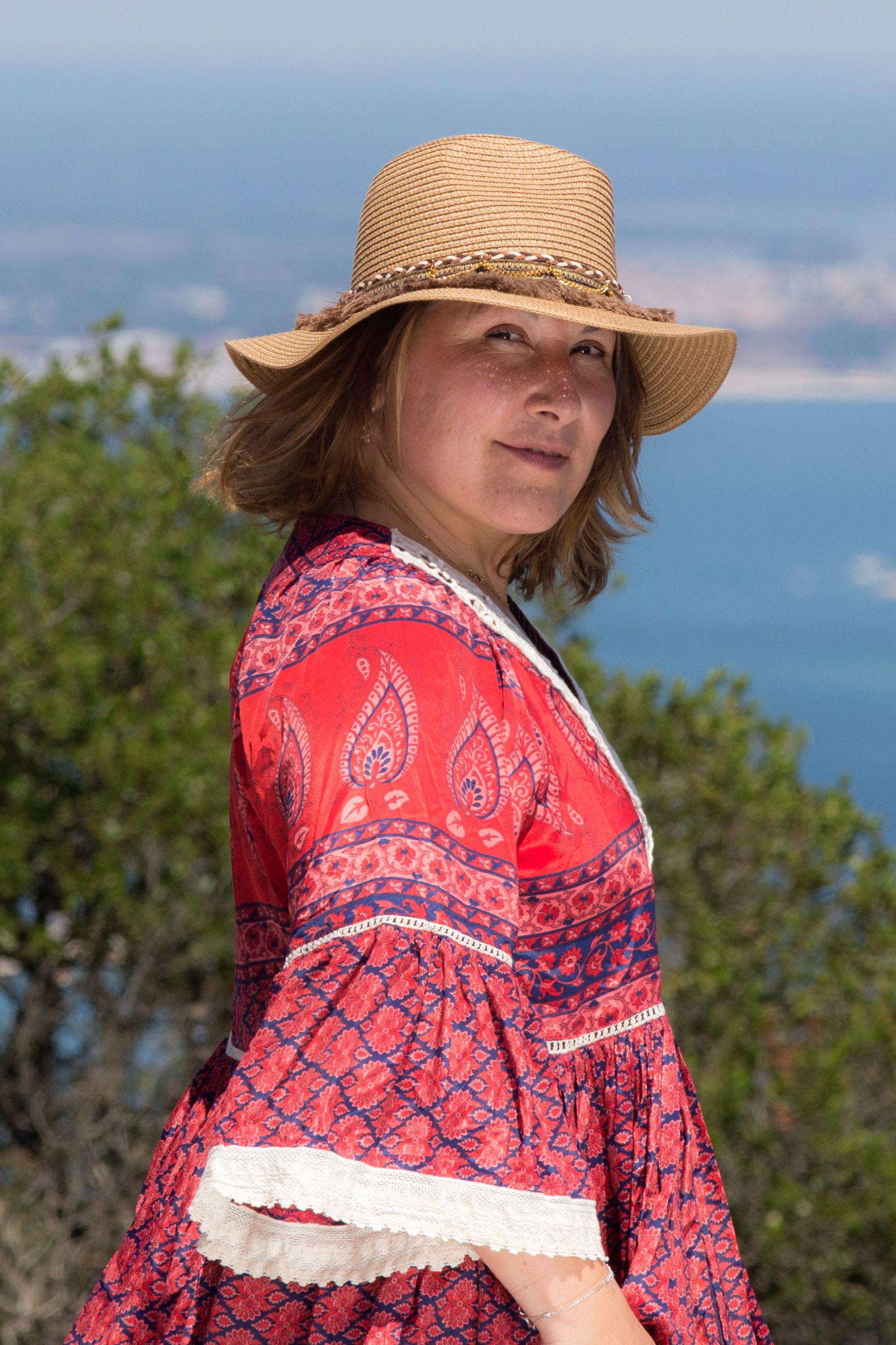 Woman in a patterned red dress with hat, enjoying a coastal viewpoint view.