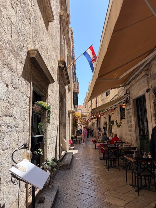 Free Picturesque street in Dubrovnik's Old Town with outdoor cafe and Croatian flag under sunny skies. Stock Photo