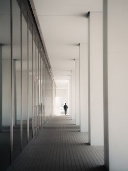 A lone individual walks through a modern building's minimalist corridor with glass walls.