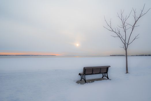 A tranquil winter scene in Montreal with snow-covered ground, a bench, a tree, and a setting sun.