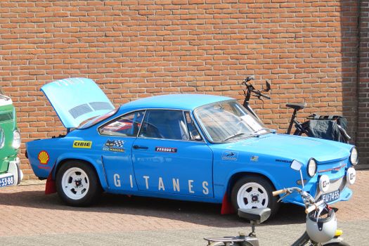 Blue vintage Gitanes race car with hood open, parked against a brick wall, showcasing classic design.