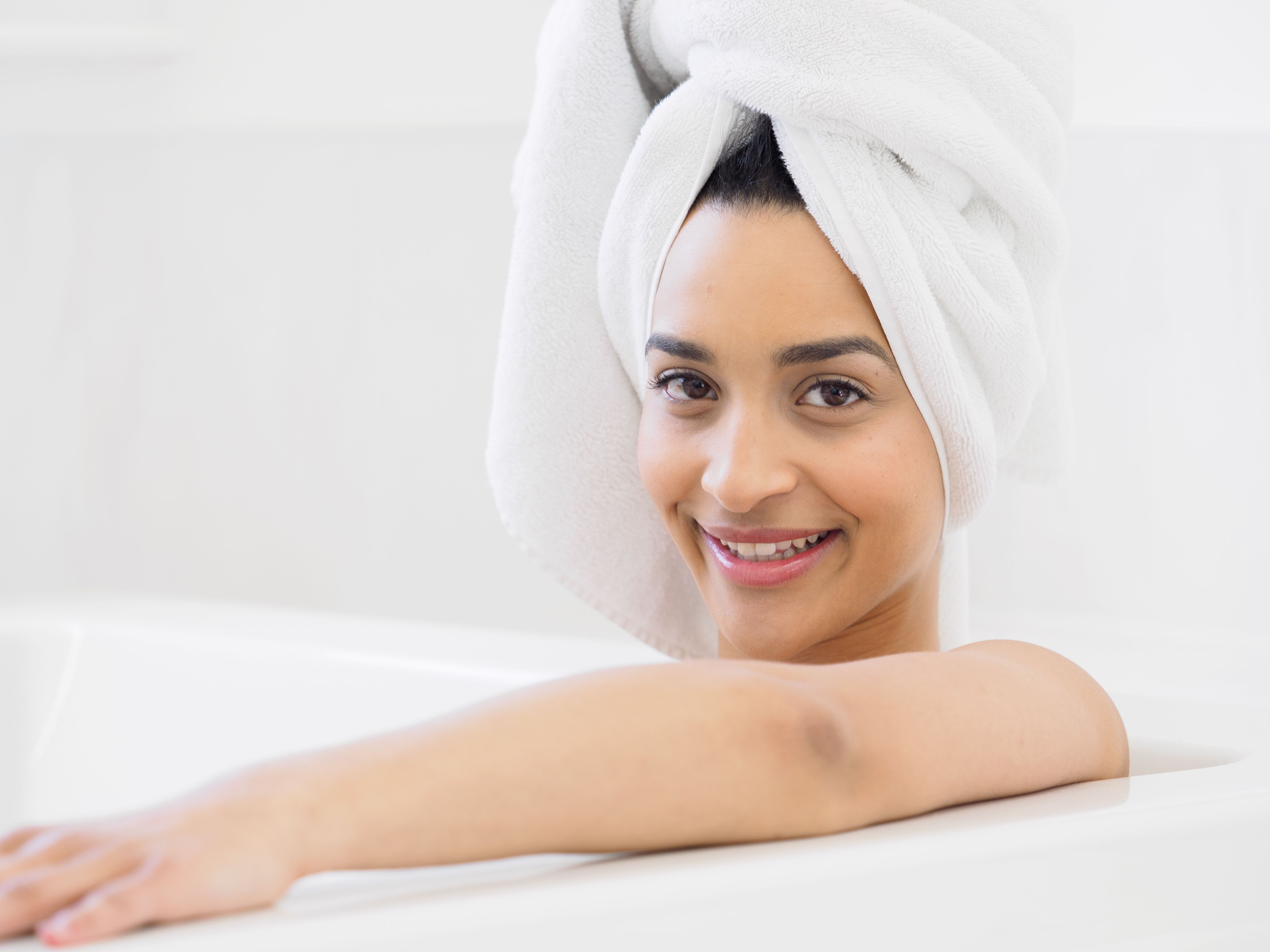 Smiling woman enjoying a relaxing bath with a towel wrapped on her head, indoors.